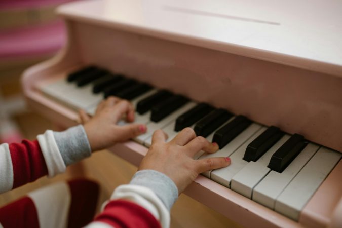 Des petits doigts d'enfant avec du vernis rose s'exercent sur les touches d'un mini piano à queue rose pastel, une scène charmante d'apprentissage pour les pianistes débutants.