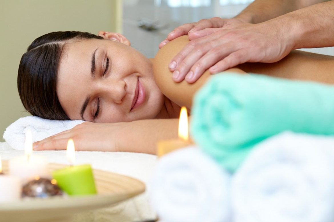Jeune femme allongée les yeux fermés recevant un massage relaxant des épaules dans un spa tamisé avec bougies et serviettes.
