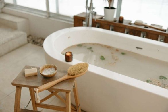 Baignoire îlot blanche remplie d'eau trouble avec des éléments naturels, accompagnée d'un petit tabouret en bois portant des accessoires de bien-être.